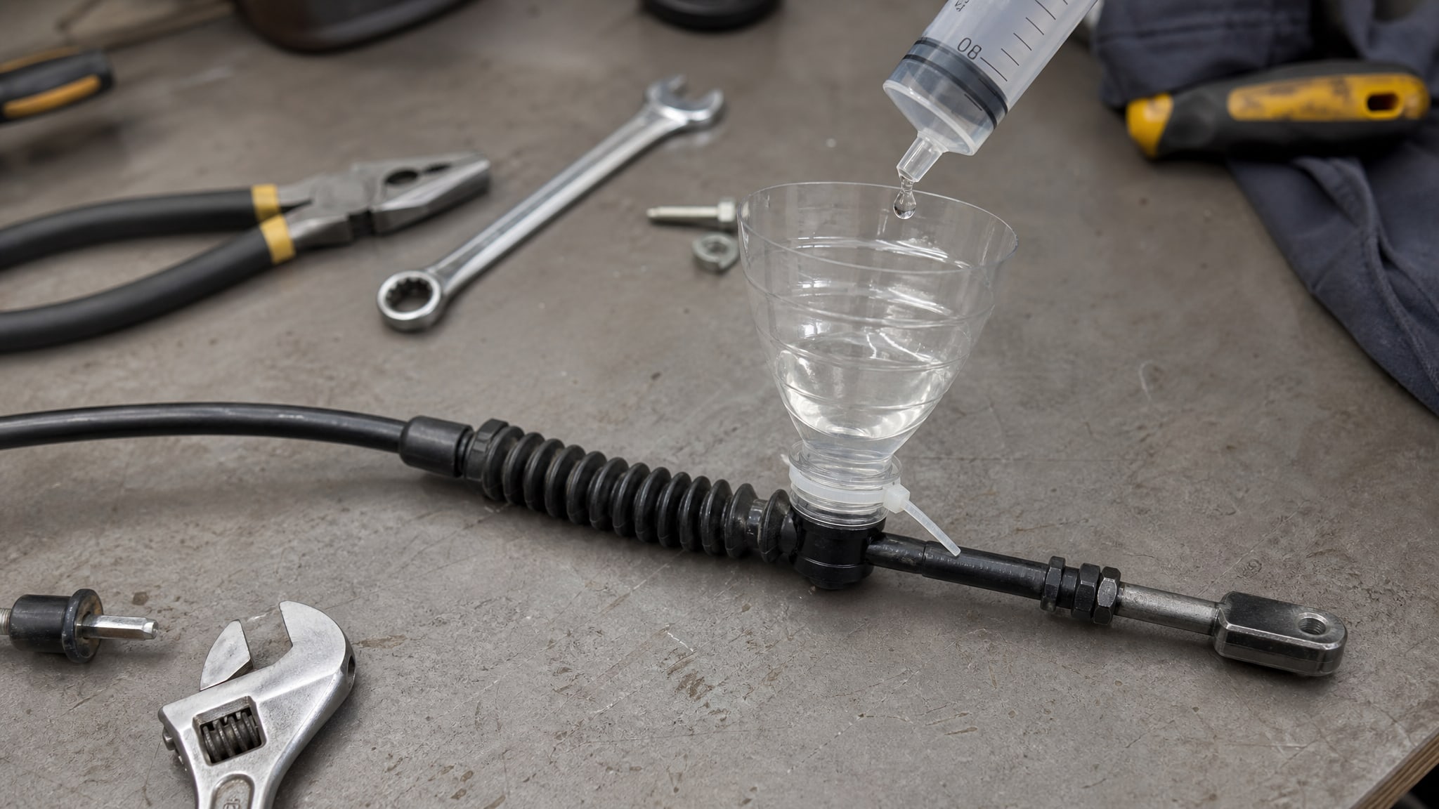 На горизонтальной поверхности лежат тросик сцепления, воронка из бутылки и шприц без иглы, из которого капает жидкость.